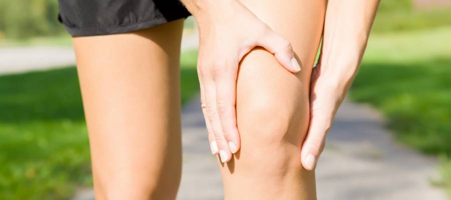 Açık havada spor kıyafeti giymiş bir kişi, sağ dizini elleriyle tutarak diz bölgesindeki ağrıyı ifade ediyor.